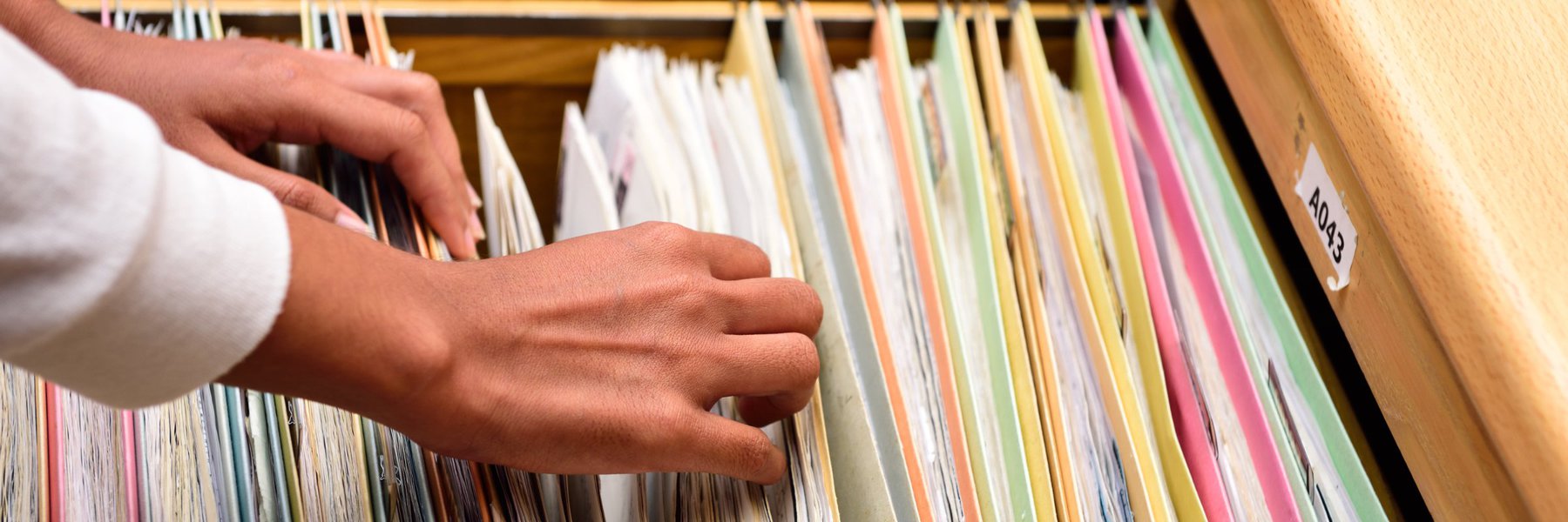 Hands are reviewing colorful files inside a filing cabinet.