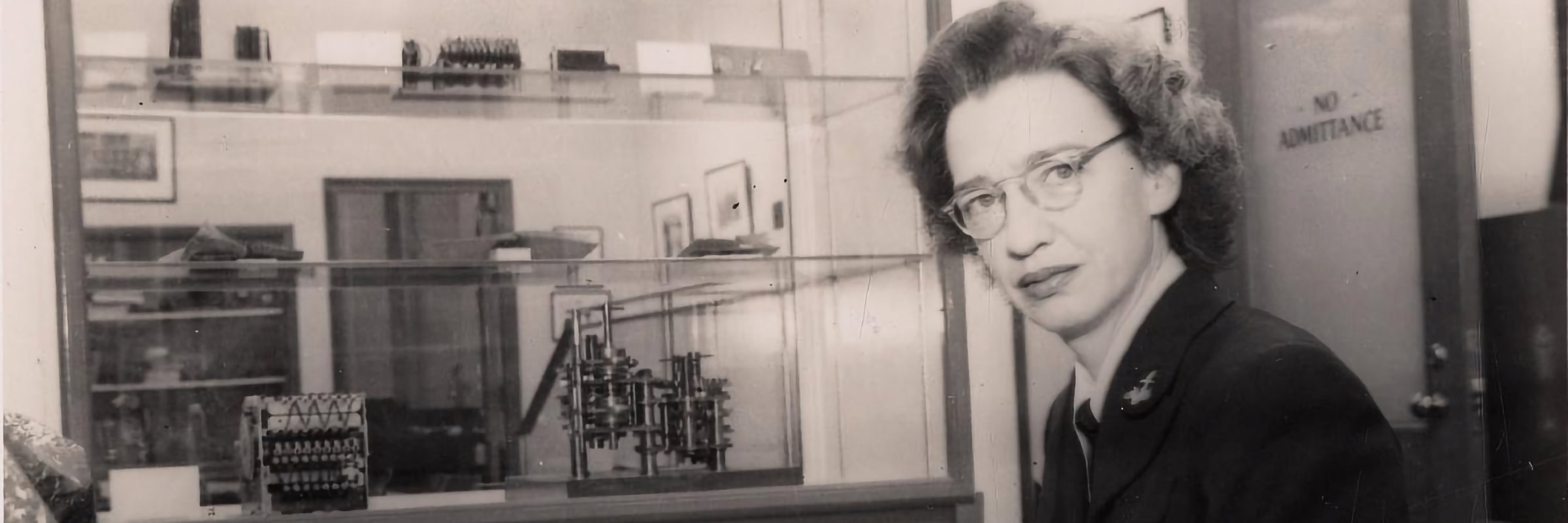 A photo of Lieutenant Grace Hopper looking at the camera from her desk, with mechanical instruments in the background.