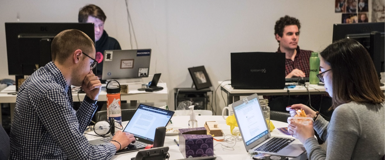 A photo of the MuckRock office, including staff sitting around a table evaluating requests with staff in the background.