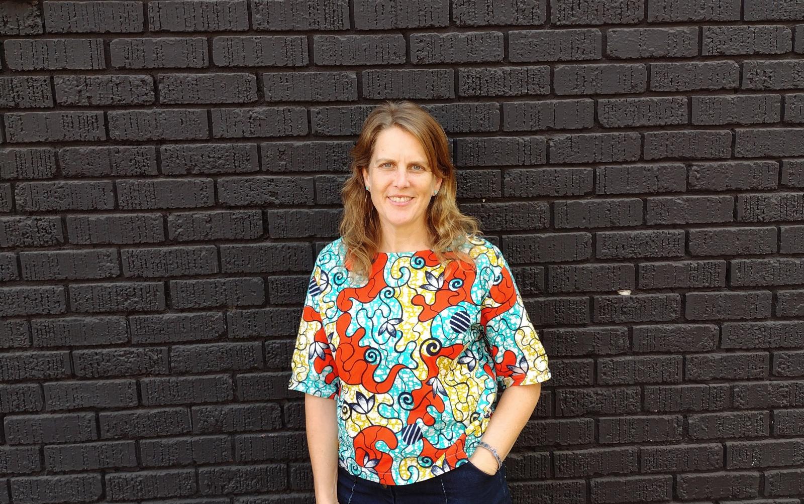 Photo of Amanda Hickman, wearing a brightly colored shirt, standing in front of a black brick wall